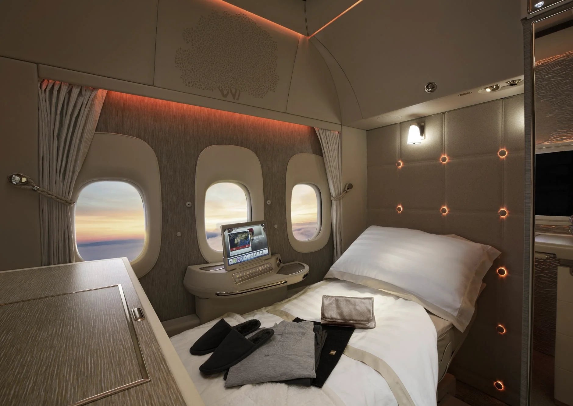 An enclosed First Class suite with a made bed and three windows onto the sunset